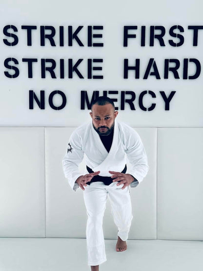 Person wearing BLITZ Sun & Moon unisex Brazilian Jiu-Jitsu Gi, posing in a dojo with motivational wall text "Strike First Strike Hard No Mercy"