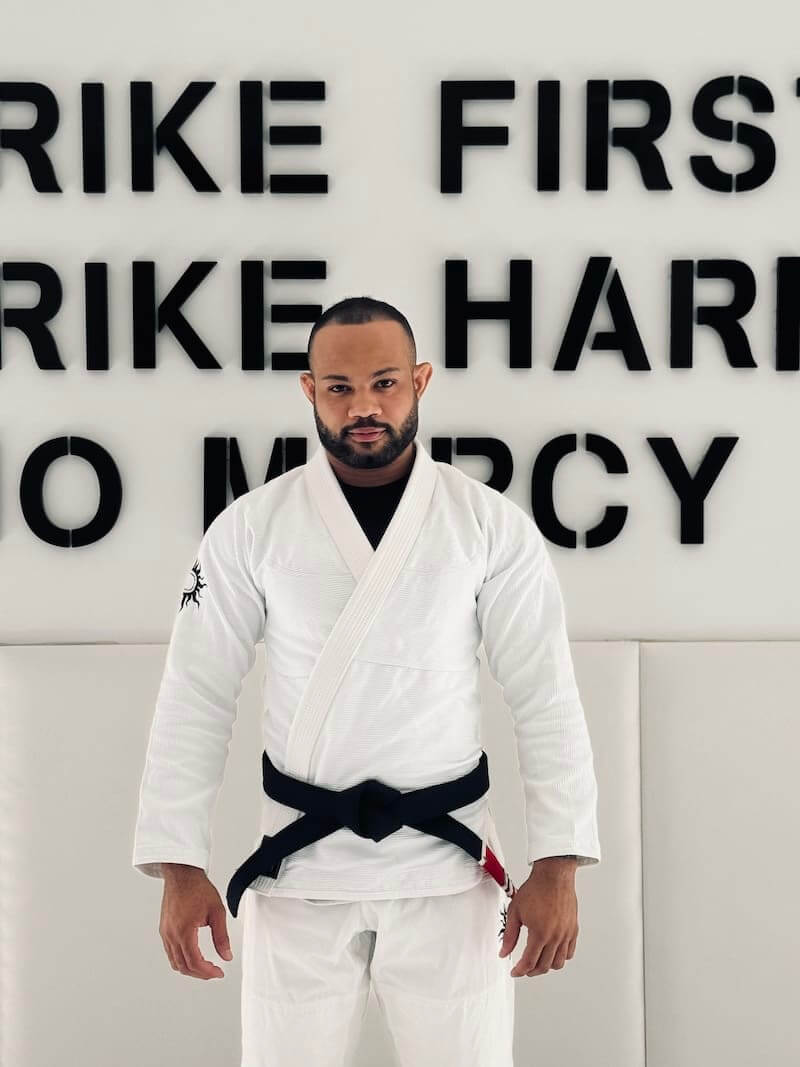 Male athlete in BLITZ Sun & Moon Brazilian Jiu-Jitsu Gi standing in front of motivational wall text at training facility.