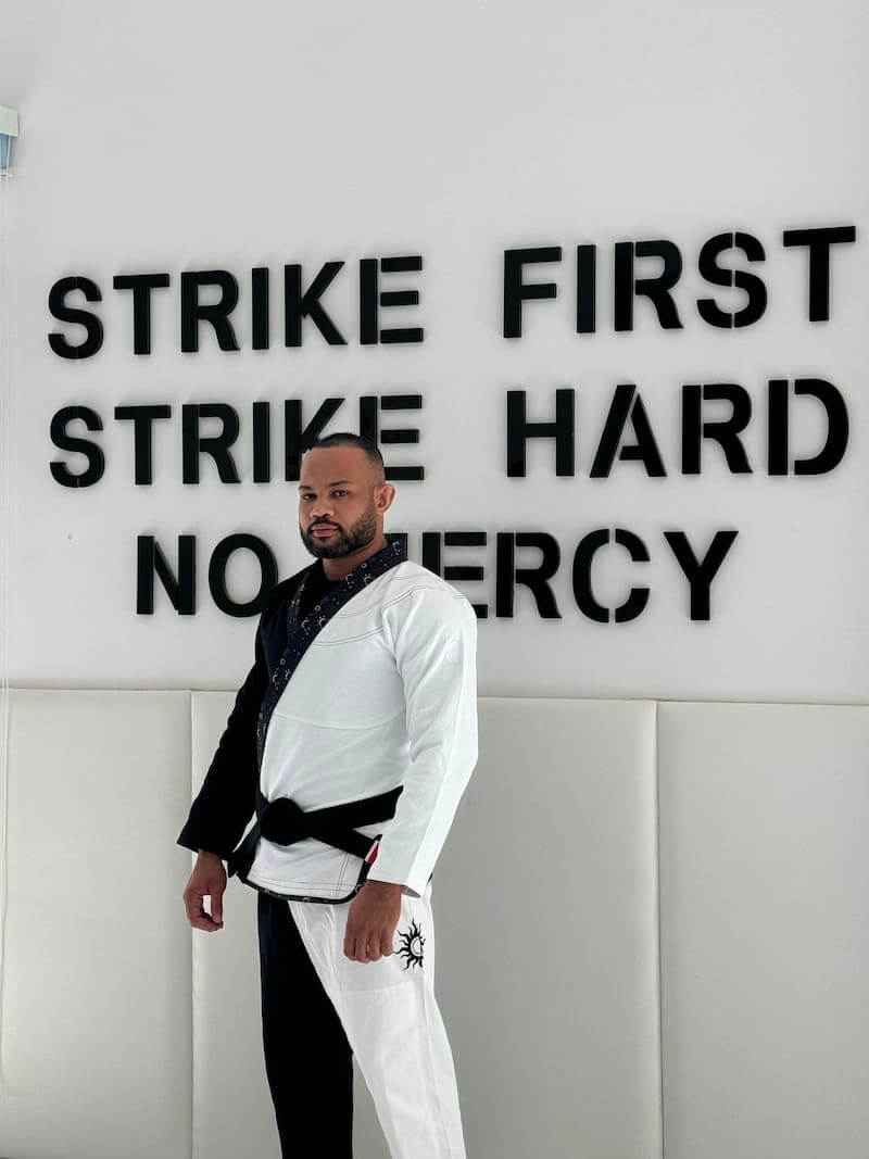 Unisex Brazilian Jiu-Jitsu Gi Model Posing in Front of "Strike First, Strike Hard, No Mercy" Wall Sign