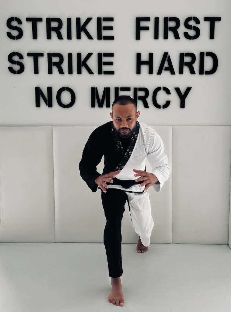 Person wearing BLITZ Sun & Moon Brazilian Jiu-Jitsu Gi in front of "Strike First, Strike Hard, No Mercy" wall sign.