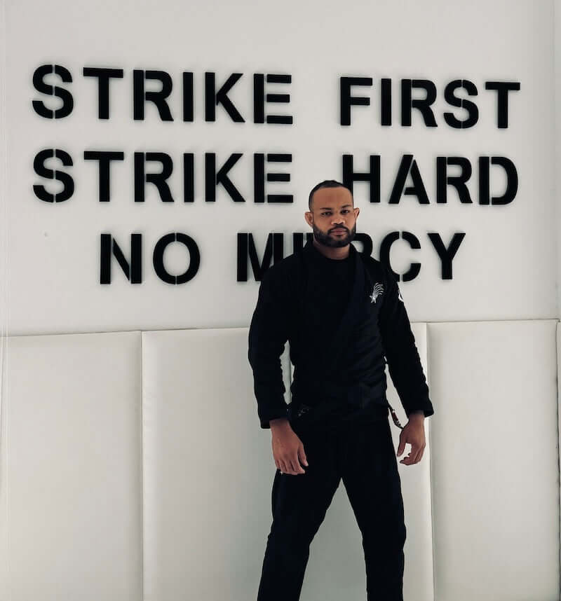 Homem vestindo o kimono de Jiu-Jitsu brasileiro BLITZ Sun & Moon em pé contra uma parede com mensagens motivacionais.