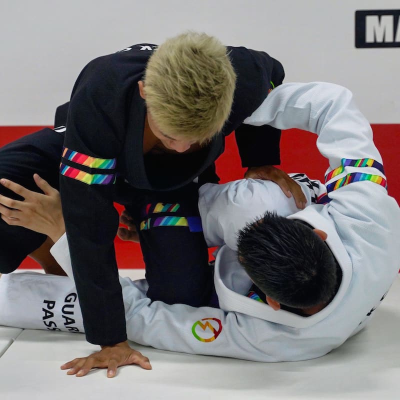 Two people practicing Brazilian Jiu-Jitsu, one in a white rainbow pattern gi and the other in a black rainbow pattern gi