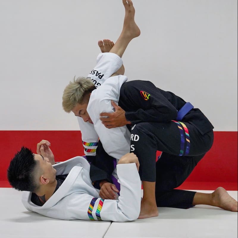 Two people practicing Brazilian Jiu-Jitsu, one in a black rainbow pattern gi and the other in a white rainbow pattern gi