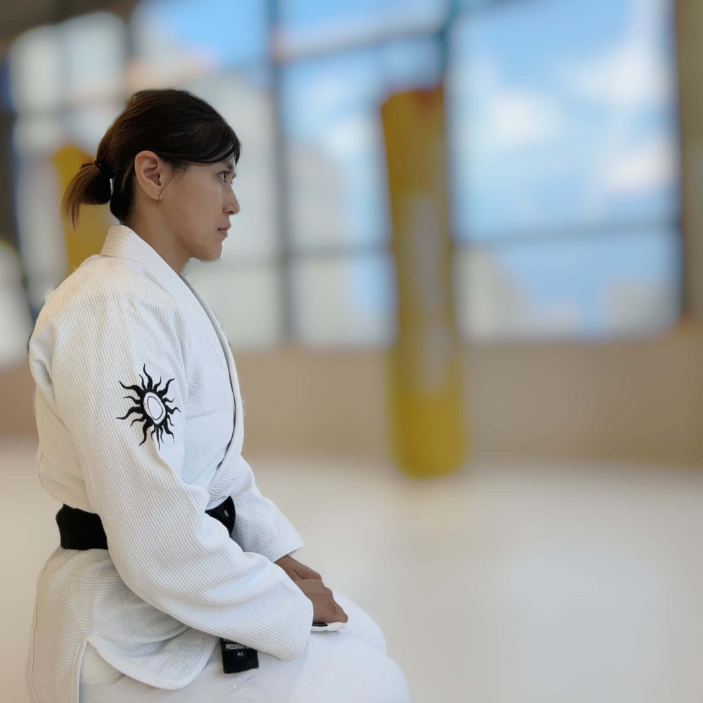 Female martial artist in BLITZ Sun & Moon Brazilian Jiu-Jitsu Gi, seated and focused in training environment.